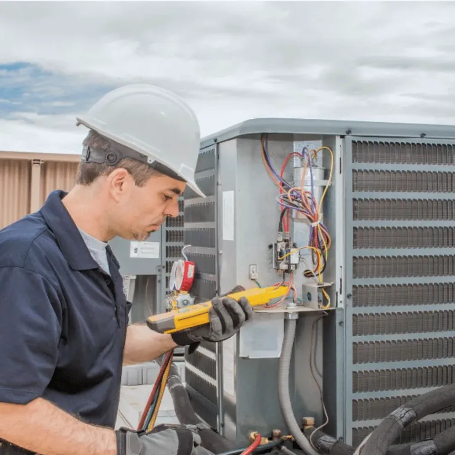 HVAC technician in a hard hat testing an outdoor air conditioner with a multimeter